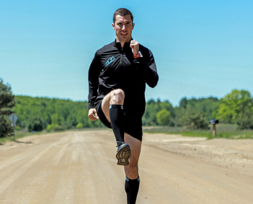 man on a bright dirt road with green trees in the background. Man is doing running drills.