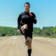 man on a bright dirt road with green trees in the background. Man is doing running drills.