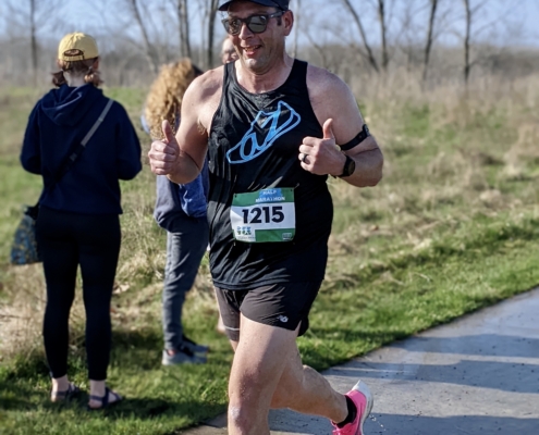 Runner racing a half marathon on a paved. He is smiling giving a thumbs up while racing.