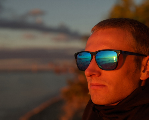 man sitting on the beach at sunset. His face is orange from the light. He is wearing sunglasses with the reflection of the water and sunset.