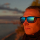 man sitting on the beach at sunset. His face is orange from the light. He is wearing sunglasses with the reflection of the water and sunset.