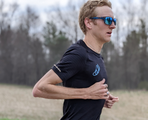 Close up of man running with sunglasses. He is wearing a tshirt. The background is a field and trees.