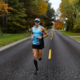 man running on the road with the fall foliage of Michigan in the background. Man running is wearing blue and a visor camp, sunglasses, and tall socks.