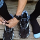 Close up of hands tying running shoes. You can see the watch on the runners wrist. The runner is wearing tall black socks with blue toes and stripes.