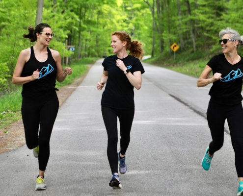 three women running together in the woods, smiling and enjoying one another.