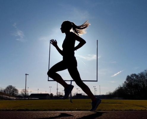 Woman running on the track with a long ponytail. She is a silhouette with blues behind her. She is in a powerful part of her stride.