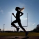 Woman running on the track with a long ponytail. She is a silhouette with blues behind her. She is in a powerful part of her stride.